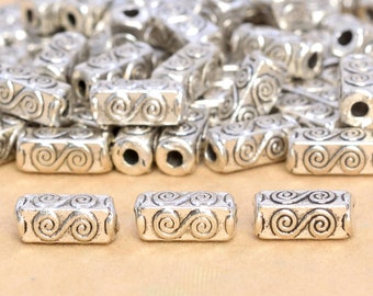 Peut inclure: Gros plan sur trois perles en métal argenté avec un motif en spirale. Les perles sont disposées en rangée sur une surface brun clair. Les perles mesurent environ 2,5 cm de long et 1,25 cm de large.