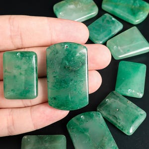 May include: A collection of green jade pendants and stones. The pendants are rectangular with rounded edges and a small hole at the top for stringing. The stones vary in shape, with some rectangular and others more geometric. The jade has a translucent quality.