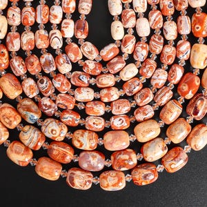 Puede incluir: Primer plano de un collar con numerosas cuentas ovaladas en tonos naranja, marrón y crema. Las cuentas están ensartadas con espaciadores transparentes y facetados, creando un efecto de capas. El collar se presenta sobre un fondo negro.