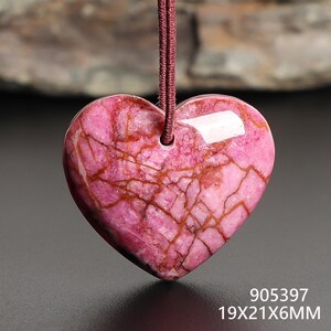 May include: A heart-shaped pendant made of pink stone with brown veining. The pendant is suspended from a maroon cord. The dimensions are 19x21x6mm.