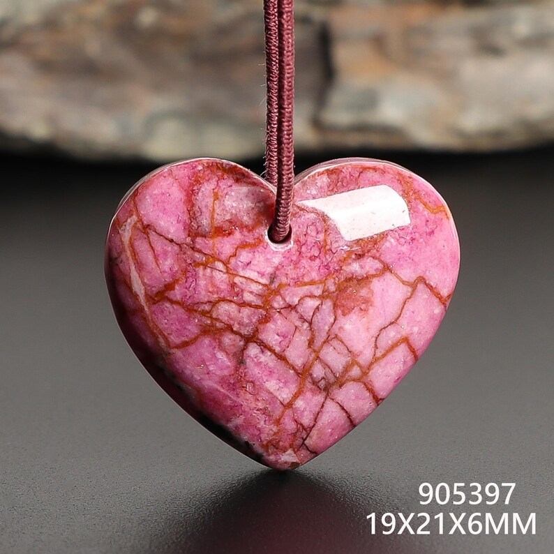 May include: A heart-shaped pendant made of pink stone with brown veining. The pendant is suspended from a maroon cord. The dimensions are 19x21x6mm.