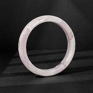 May include: A circular bangle bracelet crafted from polished stone with a translucent, pale pink and white marbled appearance. The surface features subtle variations in color and texture throughout the smooth, rounded band structure.