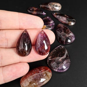 May include: Polished, teardrop-shaped gemstones in shades of purple, brown, and red. The gemstones are held in a hand, with several others scattered on a black surface. The stones have a glossy finish.