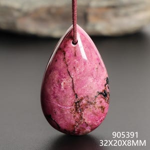 May include: A teardrop-shaped pendant with a polished, pink surface, featuring dark veining and a small black area. The pendant is suspended by a thin, reddish-brown cord. The dimensions are 32x20x8mm.