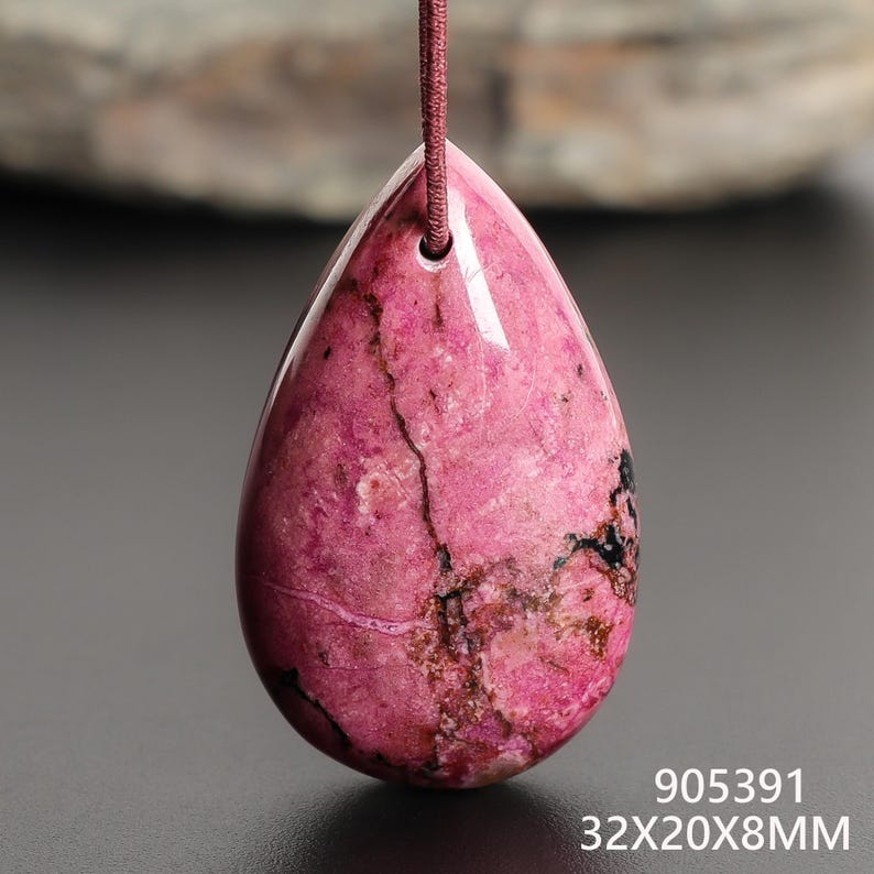 May include: A teardrop-shaped pendant with a polished, pink surface, featuring dark veining and a small black area. The pendant is suspended by a thin, reddish-brown cord. The dimensions are 32x20x8mm.