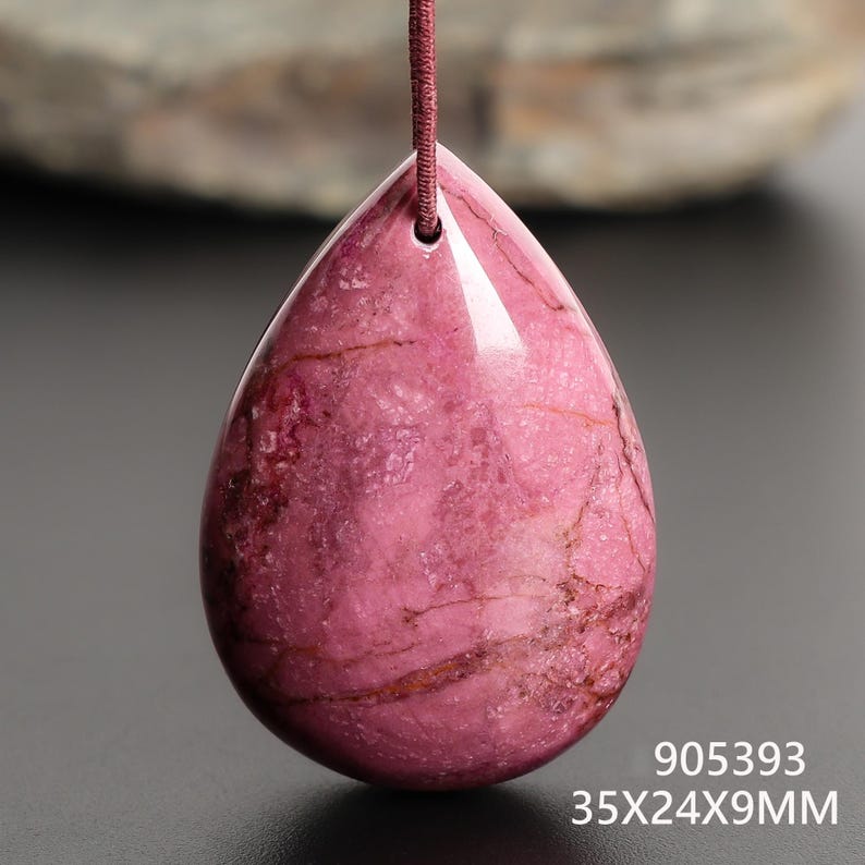 May include: A polished, teardrop-shaped pendant in shades of pink and maroon, suspended by a maroon cord. The gemstone measures approximately 1.4 x 0.9 x 0.4 inches. The pendant has a smooth, glossy surface with natural veining. The text "905393" and "35X24X9MM" are visible.