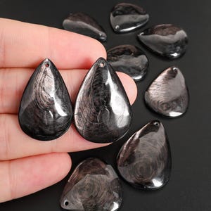 Puede incluir: Un conjunto de colgantes negros en forma de lágrima con acabado brillante. Los colgantes tienen un agujero para ensartar y presentan un patrón arremolinado y texturizado. Ideales para la creación de joyas o manualidades.