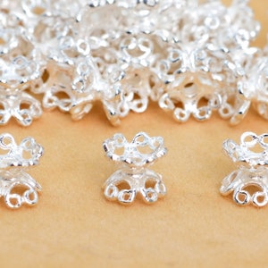 May include: Silver-toned metal bead caps with a delicate, intricate design. The caps have a round opening at the top and a flat base. They are perfect for adding a touch of elegance to jewelry projects.