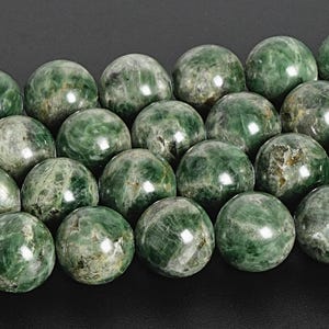 May include: Close-up of a collection of green gemstone beads. The beads are round, with a polished surface and varying shades of green and gray. The beads are approximately 1 inch in diameter.