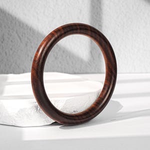 May include: A circular bangle bracelet crafted from dark brown wood with a polished finish. The wood grain features visible natural patterns and deep mahogany tones throughout the smooth, rounded surface of the solid circular jewelry piece.