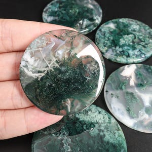 May include: A collection of polished, round moss agate stones. The stones display a mix of translucent white and green patterns, resembling moss. One stone is held in a hand, with others scattered on a dark surface. These stones are ideal for jewelry making or decorative purposes.