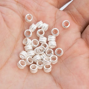 May include: A pile of silver-toned metal bead spacers, with a textured surface, in the palm of a hand.