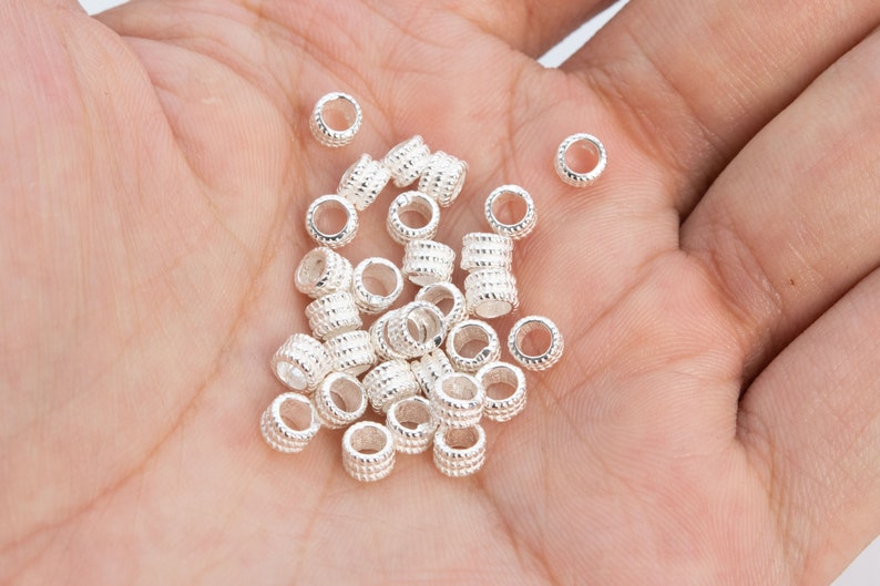 May include: A pile of silver-toned metal bead spacers, with a textured surface, in the palm of a hand.