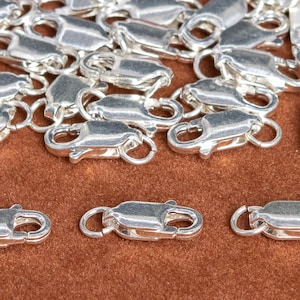 May include: A pile of silver-toned lobster clasps, a type of jewelry fastener, on a brown surface. Three clasps are separated from the pile and are positioned in a row.