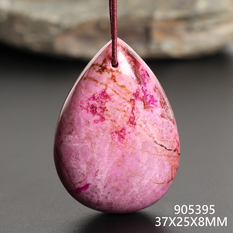 May include: A teardrop-shaped pink pendant with a brown cord. The pendant has a polished surface with darker pink and brown veining. The dimensions are 37x25x8mm.