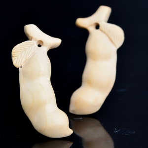 Puede incluir: Dos tallas de madera de una vaina de frijol con un detalle de hoja. Las tallas son de color marrón claro y tienen un agujero en la parte superior para colgar.