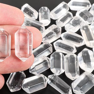 May include: Clear quartz crystal pendants with a hexagonal shape. Two pendants are held in a hand, with many more scattered in the background. The crystals are transparent and faceted, reflecting light. These pendants are suitable for jewelry making or crafting.