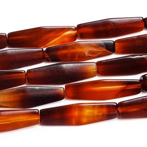 May include: Close-up of a strand of brown, oblong beads. The beads have a polished, translucent appearance with variations in color, ranging from dark brown to amber. The beads are strung together, creating a linear pattern against a white background.