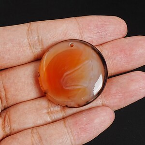 May include: A round, polished gemstone pendant with a hole for stringing. The pendant features a swirling pattern of orange, white, and brown colors. The stone is held in a hand against a black background.