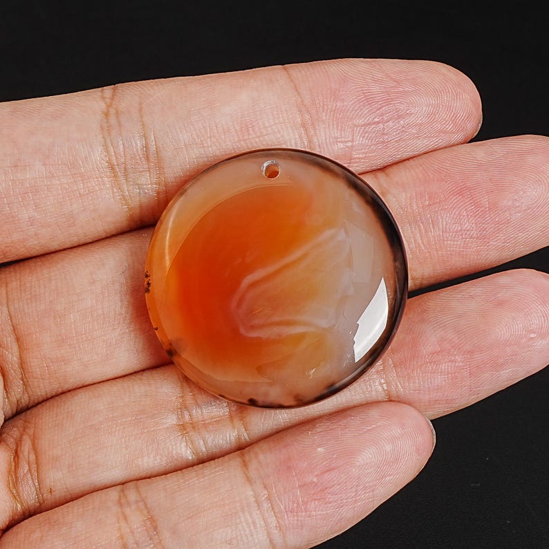 May include: A round, polished gemstone pendant with a hole for stringing. The pendant features a swirling pattern of orange, white, and brown colors. The stone is held in a hand against a black background.