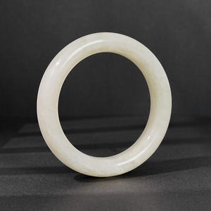 May include: A smooth, circular bangle bracelet crafted from a solid piece of off-white, translucent stone. The bangle features a polished, rounded surface with subtle variations in its pale, milky color throughout the entire circular form.