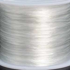 May include: A spool of clear elastic string wound around a white plastic bobbin. The string is translucent and appears to be very thin.