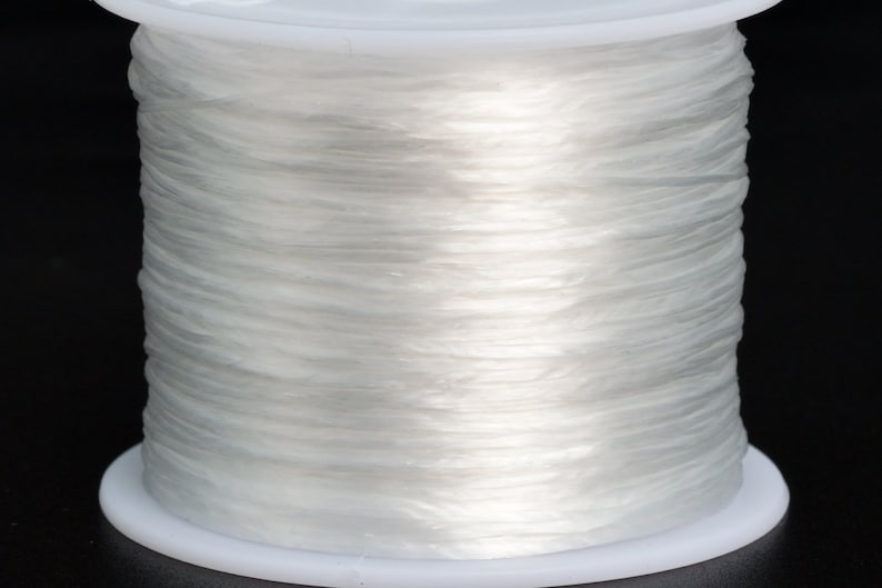 May include: A spool of clear elastic string wound around a white plastic bobbin. The string is translucent and appears to be very thin.
