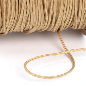 May include: A spool of tan paracord, a type of strong nylon cord, is shown on a brown cardboard spool. A length of the cord is hanging down from the spool.