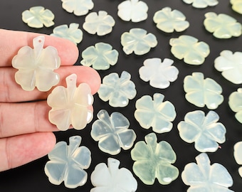 May include: A collection of pale green and white clover-shaped gemstone pendants. The pendants have a four-leaf design with a small hole at the top for stringing. The background is black.
