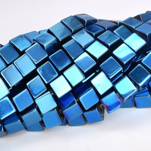 Può includere: Una collana di perline quadrate metalliche blu. Le perline sono in ematite e hanno una finitura lucida e iridescente.