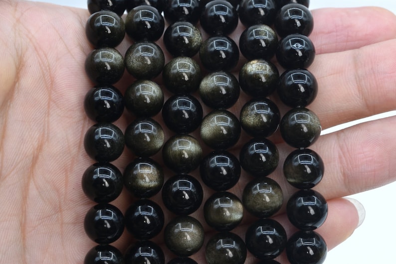 May include: A strand of black and gold gemstone beads. The beads are round and have a smooth, polished finish. The beads are strung on a clear elastic cord.