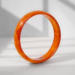 Puede incluir: Un brazalete circular de tipo esclava con un tono naranja vibrante y un patrón jaspeado en espiral por toda su superficie lisa y pulida. El accesorio presenta un perfil redondeado y un acabado brillante que refleja la luz en su estructura.