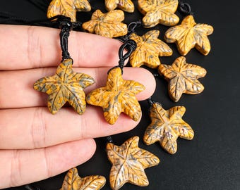 May include: Close-up of multiple maple leaf pendants. Each pendant is carved from a stone with a yellow and gray pattern. The pendants are strung on black cords. The background is black.