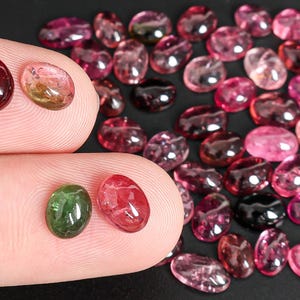 May include: A collection of oval-shaped gemstones in various shades of pink, red, and green. Some gems are held between fingers, while others are scattered on a dark surface. The gems vary in size and color saturation.
