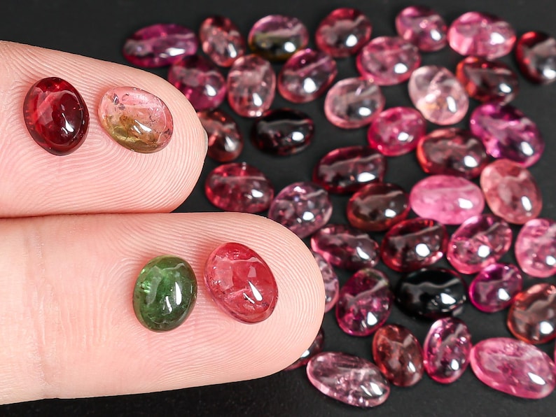 May include: A collection of oval-shaped gemstones in various shades of pink, red, and green. Some gems are held between fingers, while others are scattered on a dark surface. The gems vary in size and color saturation.