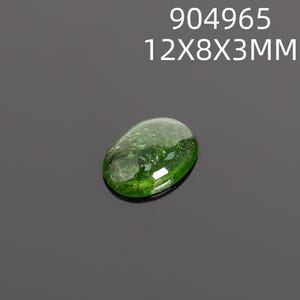 May include: A polished, oval-shaped green gemstone. The gem is a vibrant, translucent green with a glossy surface. The text "904965 12X8X3MM" is printed above the gemstone. The gemstone measures approximately 0.47 x 0.31 x 0.12 inches.
