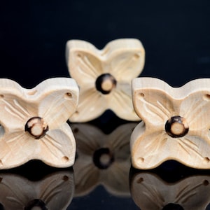 Puede incluir: Tres cuentas de madera con forma de flor con un centro marrón oscuro. Las cuentas son de color marrón claro y tienen un acabado liso y pulido.