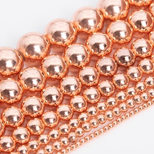 May include: A collection of rose gold beads in various sizes, arranged in rows from largest to smallest. The beads are smooth and shiny, with a metallic finish.