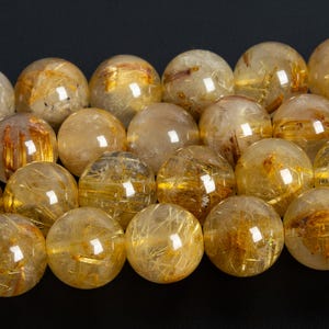 May include: A close-up shot of a collection of golden rutile quartz spheres. The translucent spheres have internal golden needle-like inclusions, creating a shimmering effect. The spheres are arranged closely together against a dark background.