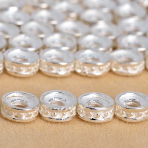 May include: Silver-toned metal beads with a decorative design. The beads are round and have a hole in the center. They are arranged in a row on a brown surface.