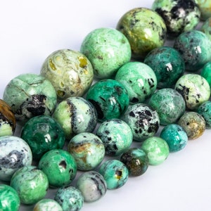 May include: Strands of round gemstone beads in shades of green, teal, and brown. The beads vary in size and have a marbled appearance, with darker spots and patterns. The beads are arranged on a white surface.