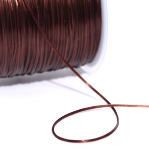 Może przedstawiać: Szpula ciemnobrązowego elastycznego sznurka. Sznurek jest ciasno nawinięty na białą plastikową szpulę. Kawałek sznurka wystaje ze szpuli, tworząc pętlę. Tło jest białe.