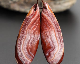 May include: Two teardrop-shaped pendants with a natural stone appearance. The pendants feature a layered pattern of red, white, and brown hues, with a polished surface. They are strung together with a brown cord, suitable for jewelry making.