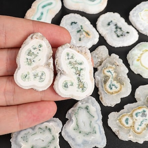 May include: A collection of polished agate slices with a variety of patterns and colors. The slices are primarily white with green, blue, and brown inclusions. Two slices are held in a hand.