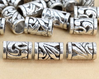 May include: Silver metal hair beads with an intricate, ornate design. The beads are cylindrical and have a hollow centre.
