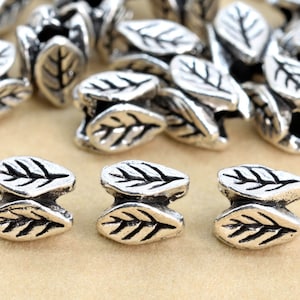 May include: Silver tone metal leaf shaped beads, each bead is two leaves joined together, the leaves have a detailed vein pattern.