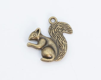 Puede incluir: Un charm de ardilla de color bronce con una cola texturizada y un lazo para sujetarlo a un collar o pulsera. La ardilla está de pie, mirando hacia la izquierda, con los brazos ligeramente extendidos. Sobre un fondo blanco.