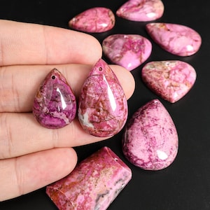 May include: A collection of pink and magenta gemstone pendants and cabochons. The teardrop and heart-shaped stones have a marbled appearance with variations in color. Some stones have holes for stringing.