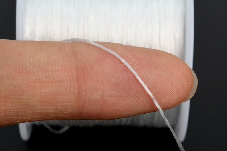 May include: A close-up of white elastic cord wound around a spool. The cord is thin and flexible, and it is being held in place by a finger.