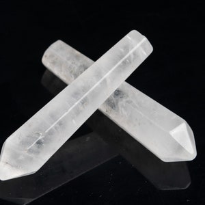 May include: Two clear quartz crystal wands, each with a pointed tip, are lying on a black surface. The wands are crossed over each other.